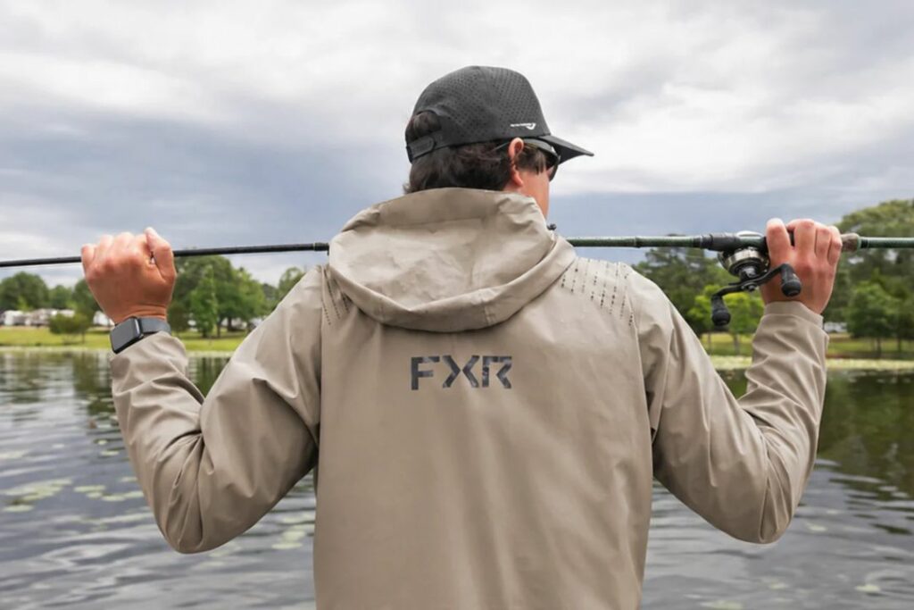 un pêcheur portant un manteau de pêche FXR tient une canne à pêche devant un cours d'eau